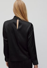 Blusa nera con maniche lunghe, texture satinata liscia, colletto alto e dettaglio a apertura a chiave sul retro fissato con un bottone circolare.