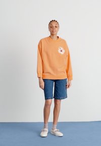 Sweat-shirt orange avec un motif floral sur la poitrine gauche, associé à un short en denim. Le mannequin porte des chaussettes grises et des chaussures claires. Fond uni.