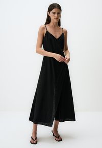 Zwarte, mouwloze maxi jurk met een V-hals, gemaakt van lichtgewicht stof. Kenmerkt een wikkelontwerp en een zijsplit voor extra detail.