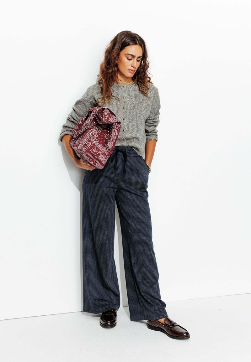 Pull gris côtelé avec boutons, pantalon large à carreaux bleu marine, et sac à motifs bordeaux, complété par des mocassins foncés.