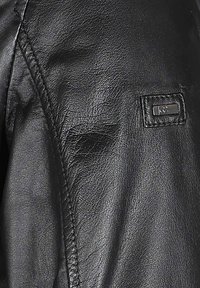 Zwarte leren jas met een gladde textuur, opvallende stiksels en een klein merklabel op de mouw. Close-up van de schouder.