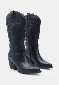 Černé kožené cowboy boty z umělé kůže s špičatými prsty, terčíkovanými květinovými vzory a bočními zipy. Blokové podpatky a dekorativní prošití po celém povrchu.