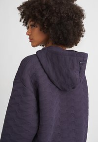 Fioletowa bluza z kapturem o teksturowanym wzorze przypominającym smugi, z kapturem i dyskretną metką na plecach. Miękka tkanina, luźny fason.