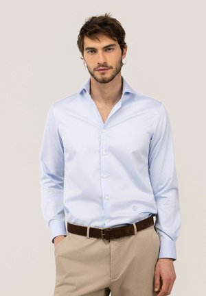 Hombre con camisa azul claro abotonada metida en pantalones beige con cinturón marrón, de pie con las manos en los bolsillos contra un fondo liso.