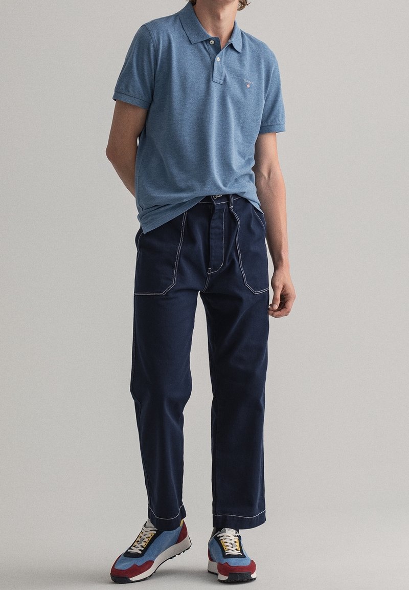 Homme portant un polo bleu, un pantalon bleu foncé avec des coutures blanches et des baskets multicolores, debout devant un fond uni.