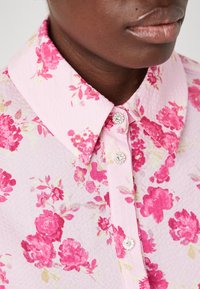 Camicia floreale rosa con tessuto testurizzato, colletto a punta e dettagli con bottoni in cristallo, caratterizzata da vivaci motivi di fiori rosa e giallo chiaro.