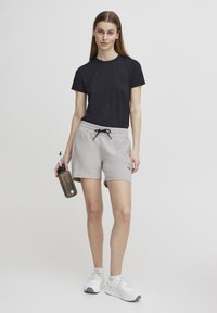 Svart atletisk t-skjorte i lett stoff; lysegrå shorts med elastisk midje og snor; holder en brun vannflaske; hvite joggesko.