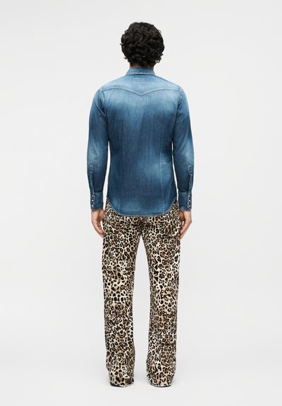 Άτομο με σγουρά μαλλιά που φοράει ξεθωριασμένο μπλε τζιν πουκάμισο και φαρδιά παντελόνια με λεοπάρ σχέδιο, στέκεται με την πλάτη σε απλό φόντο.