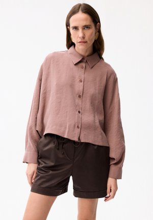 Kobieta ubrana w oversize'ową, zakurzoną różową koszulę zapinaną na guziki i ciemnobrązowe szorty ze sznurkiem, stojąca na jednolitym białym tle.