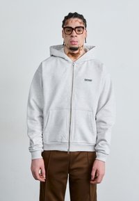 Grå zip-up hættejakke med en frontlomme; har et lille sort logo på brystet. Glat tekstur; parret med brune bukser.