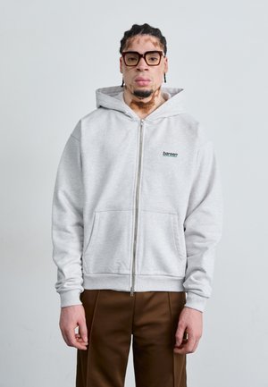 Grå zip-up hættejakke med en frontlomme; har et lille sort logo på brystet. Glat tekstur; parret med brune bukser.