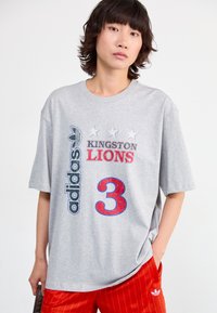 Pilkas oversize marškinėliai su spausdintu tekstu „KINGSTON LIONS“ ir numeriu „3“ raudona ir mėlyna spalvomis, su Adidas logotipu ir trimis žvaigždutėmis viršuje.