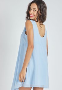 Joven mujer con cabello rizado sonriendo por encima del hombro, vestida con un vestido sin mangas de color azul claro con espalda baja.