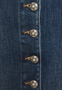 Närbild av blå denimtyg med fyra utsmyckade knappar i silvermetall som sys med orange tråd i en vertikal rad.