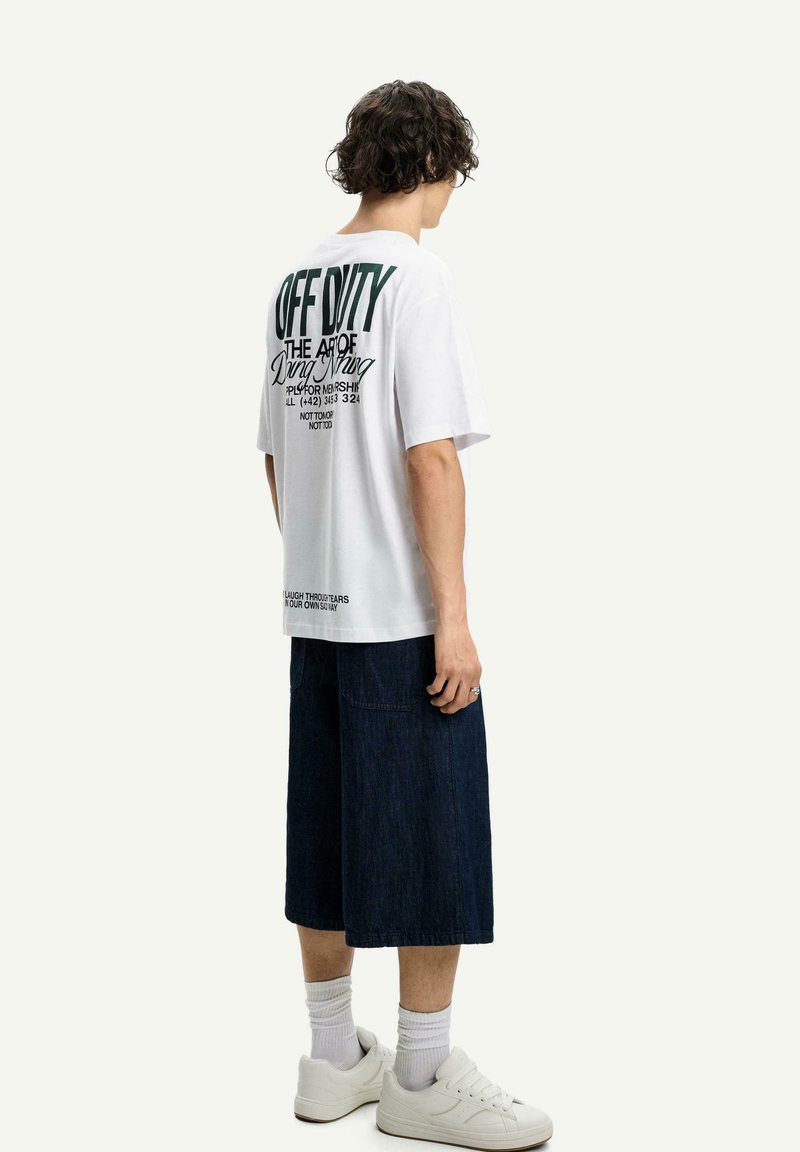 White oversized T-shirt featuring black graphic text on the back, paired with dark blue denim shorts and white sneakers.