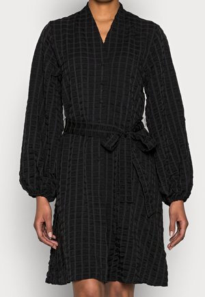 Nainen, joka pukeutuu pitkähihaiseen mustaan teksturoituun mekkoon, jossa on solmittava vyö ja napillinen edusta, seisoo yksiväristä taustaa vasten.