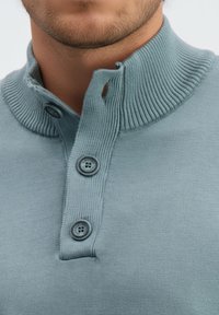 Šviesiai mėlynas megztinis su austa apykakle, trimis mygtukais ant kaklo ir lygiu paviršiumi. Artimas drabužio vaizdas.