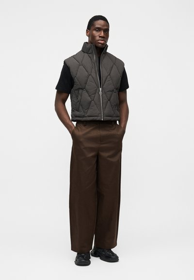 Colete acolchoado cinza escuro com gola alta, fecho de correr frontal, usado sobre t-shirt preta e calças largas castanhas, complementado por ténis pretos.