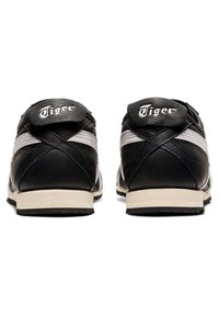 Mustat ja harmaat urheilukengät, joissa on teksturoitu nahkapinta, valkoinen logo, kontrastiväriset tikkausompelut ja litteät kumipohjat.