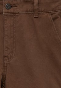 Gros plan de la poche d'un pantalon en denim marron avec passant de ceinture et bouton métal portant l'inscription "Denim Pull&Bear".