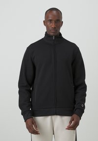 Czarna kurtka na zamek wykonana z miękkiego materiału, z wysokim kołnierzem, bocznymi kieszeniami i haftowanym logo na piersi. Prosty, zorganizowany design.