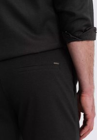 Schwarze Hose mit glatter Textur, ausgestattet mit einer dezenten Gesäßtasche und subtilen Hardware-Akzenten. Hochgekrempelter Ärmel eines dunklen Hemdes sichtbar.