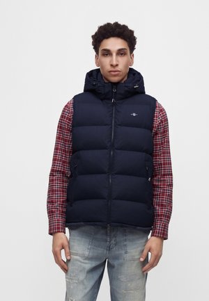 Junger Mann trägt eine marineblaue gesteppte Weste über einem roten karierten Hemd und hellblaue, abgenutzte Jeans, steht vor einem einfarbigen Hintergrund.
