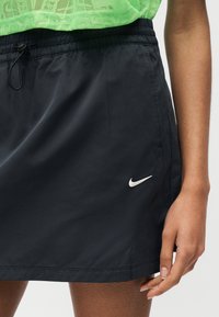 Κοντινή λήψη προσώπου που φοράει μαύρη φούστα Nike με ελαστική μέση και λευκό λογότυπο Nike swoosh, σε συνδυασμό με φωτεινό πράσινο υφασμάτινο τοπ.