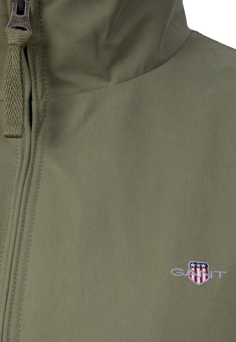 Giacca verde oliva con cerniera e logo GANT ricamato sul petto, raffigurante uno scudo con stelle e strisce.