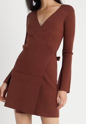 Bruine wikkeljurk met lange mouwen, V-hals en zijstrikdetail, gedragen door een persoon met donker haar en bloemenoorbellen.