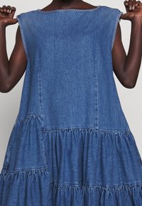 Džínová šaty s volným střihem, s kulatým výstřihem, bez rukávů a s tiered volánkovým lemem. Viditelná textura světle modrého praní.