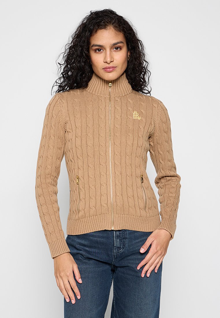 Beżowy sweter z dzianiny w warkocze z wysokim kołnierzem, złotym zamkiem błyskawicznym i dwiema bocznymi kieszeniami. Posiada małe logo na lewej piersi.