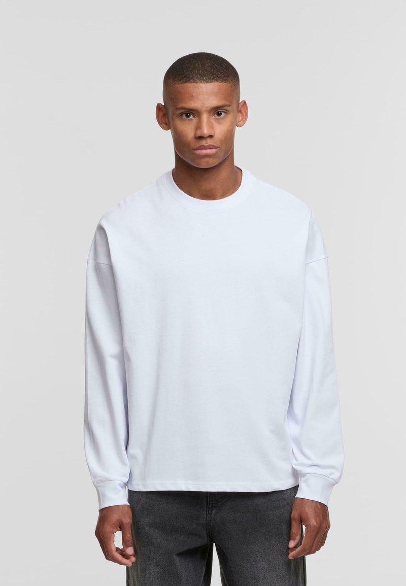 White long-sleeve T-shirt with a round neck, made of a smooth cotton fabric. Loose fit and ribbed cuffs, paired with dark gray jeans.