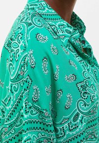 Green shirt made of lightweight fabric, featuring a paisley pattern in black and white, with a collar and button detail.