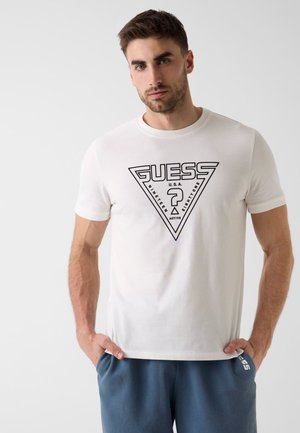 Homme portant un t-shirt blanc à logo GUESS et un short bleu, debout les mains dans les poches sur un fond uni et clair.