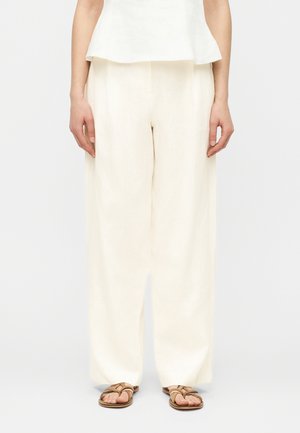 Kobieta ubrana w luźne, lniane spodnie z wysokim stanem w kolorze ecru, w połączeniu z brązowymi płaskimi sandałami, stojąca na tle jednolitej białej ściany.