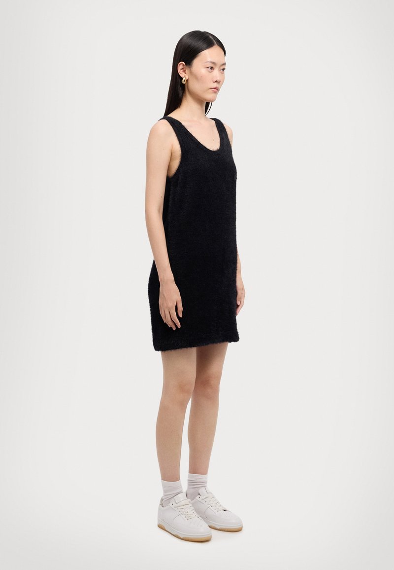 Black, sleeveless dress made of soft, textured material, with a scoop neckline and a straight silhouette, worn with white sneakers.