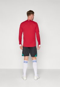 T-shirt de sport rouge à manches longues avec des poignets de manches blancs, associé à un short noir avec des accents teal, et des chaussettes blanches avec des rayures bleues.
