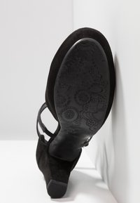 Talons en daim noir avec une semelle en caoutchouc texturée présentant des motifs floraux. La chaussure est dotée d'une bride pour un soutien supplémentaire.