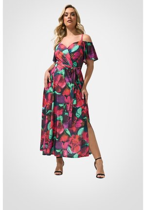 Kvinde iført off-shoulder, bælteblomstret maxikjole med røde, grønne og lilla toner, kombineret med sorte ankelstroppede hæle, stående mod hvid baggrund.