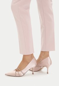 Noge u blijedo ružičastim satenskim cipelama špičastih vrhova s naramenicama ukrašenim kristalima, uparene s odgovarajućim svijetloružičastim krojenim hlačama.