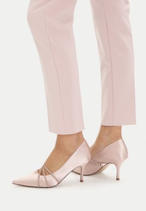 Stopy w różowych, satynowych szpilkach z odkrytym noskiem i paskami z kryształków, zestawione z dopasowanymi jasnoróżowymi spodniami na miarę.