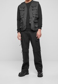 Black cargo vest with multiple front pockets, worn over a black long-sleeve shirt and matching cargo pants. Durable fabric and relaxed fit.