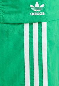 Green fabric sportswear featuring white three-stripe accents and an embroidered logo. Material appears lightweight with a smooth texture.