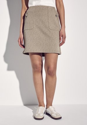 Woman wearing a beige knee-length skirt with buttoned pockets and white sneakers, standing against a plain light background.