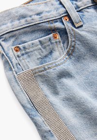 Poche de jean en denim bleu clair avec des rivets en cuivre et une bande de petits strass argentés le long de la couture extérieure.