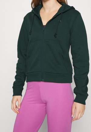 Femme portant un sweat à capuche zippé vert foncé avec cordons et des leggings rose vif, debout devant un fond clair uni.