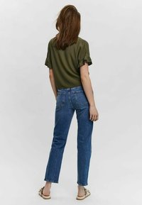 Femme de dos portant un blouse à manches courtes olive, un jean droit bleu et des sandales beiges avec des nœuds.