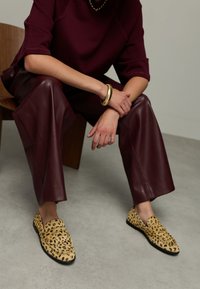 Person seated, wearing burgundy leather pants, matching top, gold jewelry, and leopard-print loafers on a gray floor.