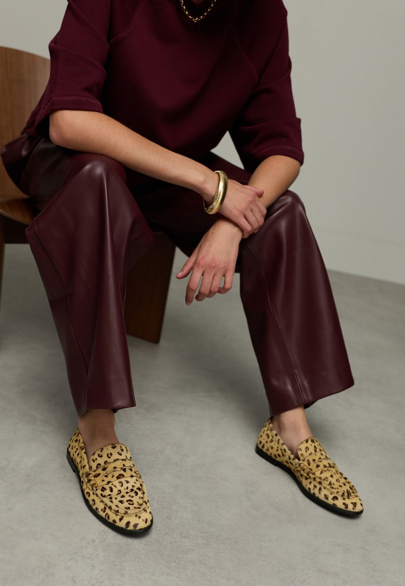 Person seated, wearing burgundy leather pants, matching top, gold jewelry, and leopard-print loafers on a gray floor.